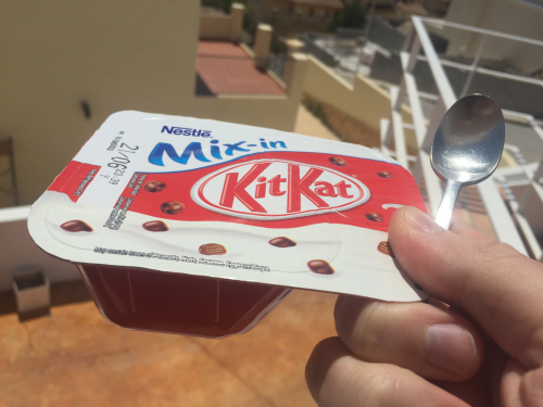 En hånd holder en uåbnet Nestlé Mix-in dessert med KitKat smag, ledsaget af en metalske. Baggrunden viser en solrig udendørsindstilling med bygninger.