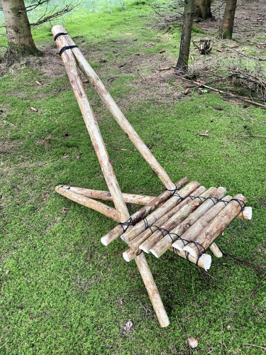 En håndlavet træstol med et rudimentært design bestående af sammenbundne grene på en grøn græsplæne.