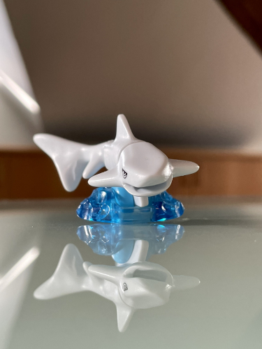 En hvid plastik hajfigur står på en gennemsigtig blå base, hvilket skaber en spejling på den blanke overflade under den.