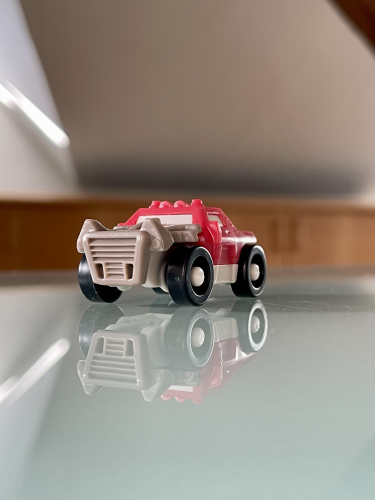 En lille plastiklegetøjsbil i rød og hvid, med markante sorte hjul, reflekteres på en skinnende overflade under indendørs belysning.